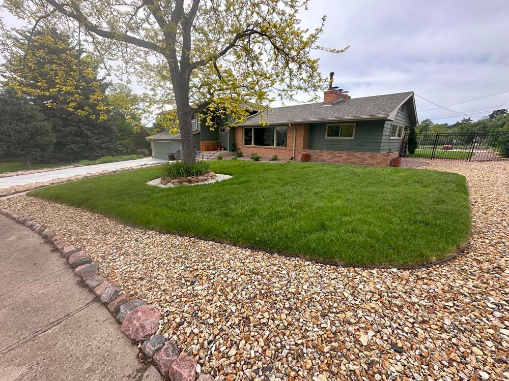 House with green siding and lawn, beige gravel border, tree, and driveway under a cloudy sky.