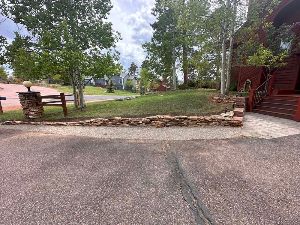 Stone wall bordering a gravel driveway and green lawn with trees and a red building.