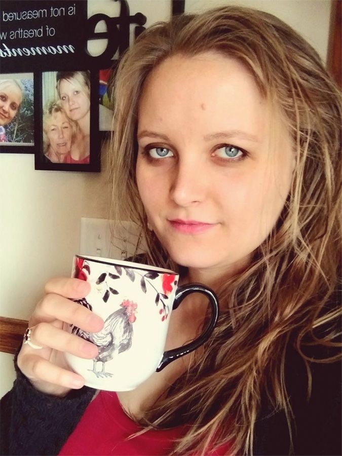 Woman with blue eyes holding a mug with a rooster design, in front of framed photos.