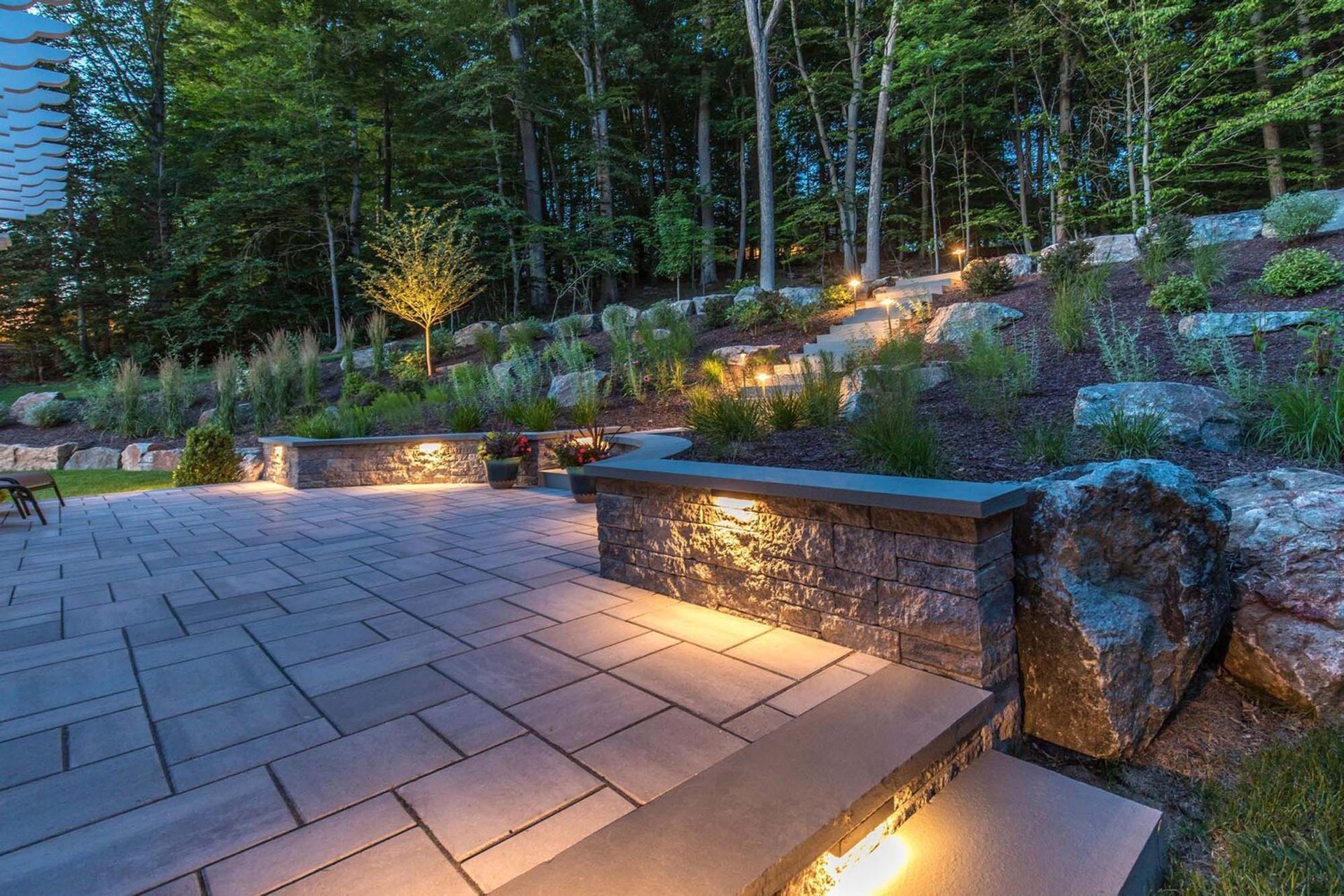 Patio with stone pavers, retaining walls, and uplighting in a wooded backyard at dusk.