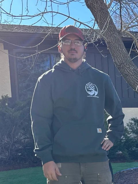 Man in a green hoodie and a red hat stands outside a house, hand on hip.