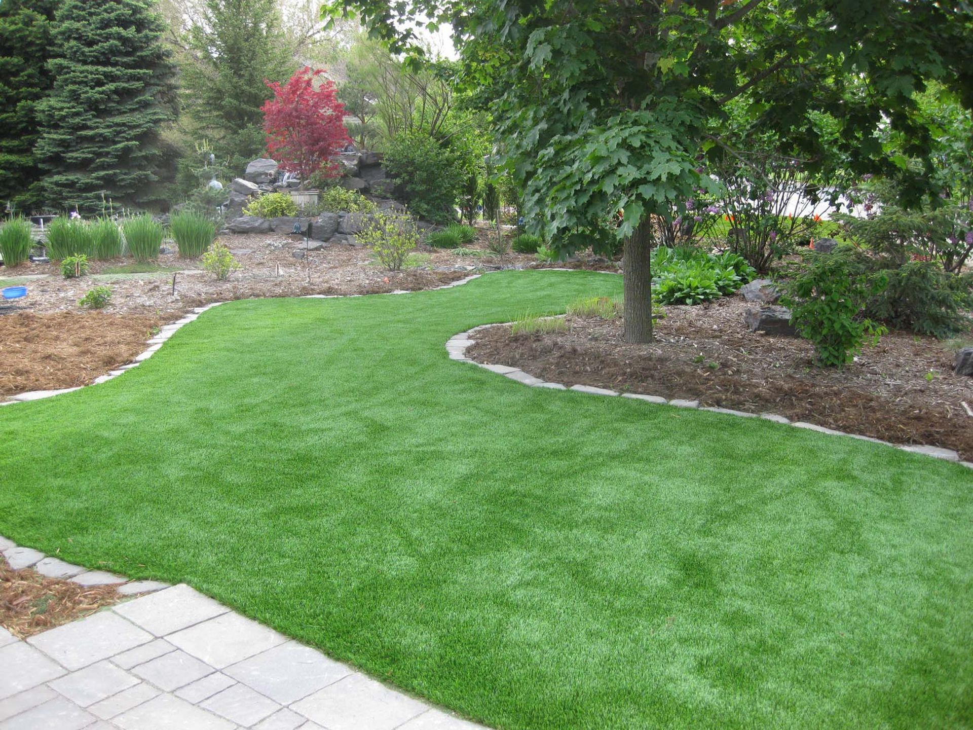 Green artificial turf pathway in a landscaped yard, bordered by stone and flowerbeds. Trees and plants surround.