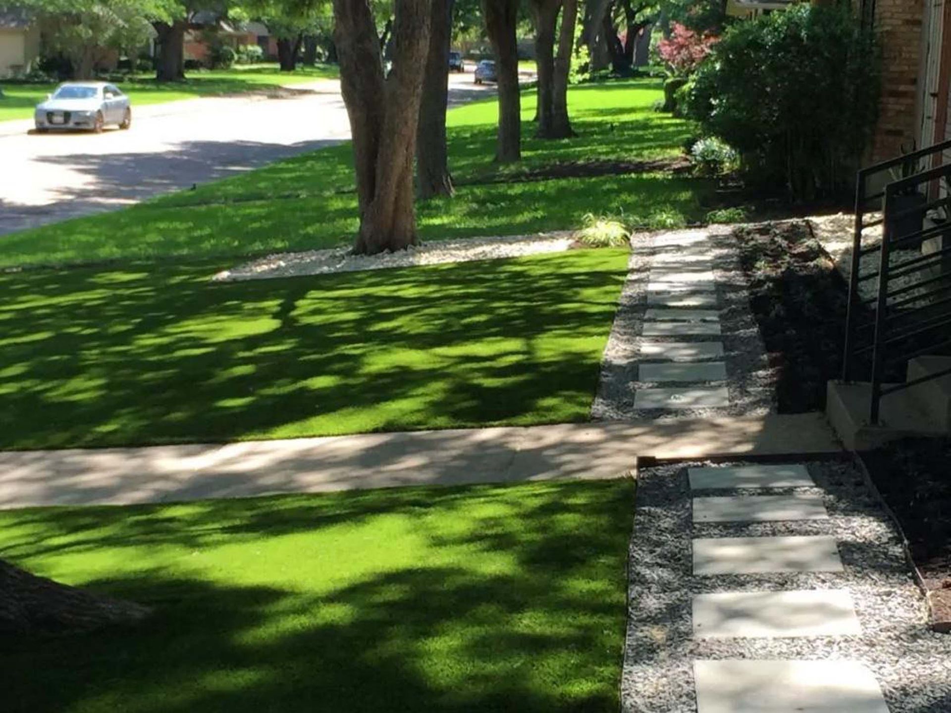 A bright green lawn with trees, a paved sidewalk, and stone pathway leading to a house entrance.