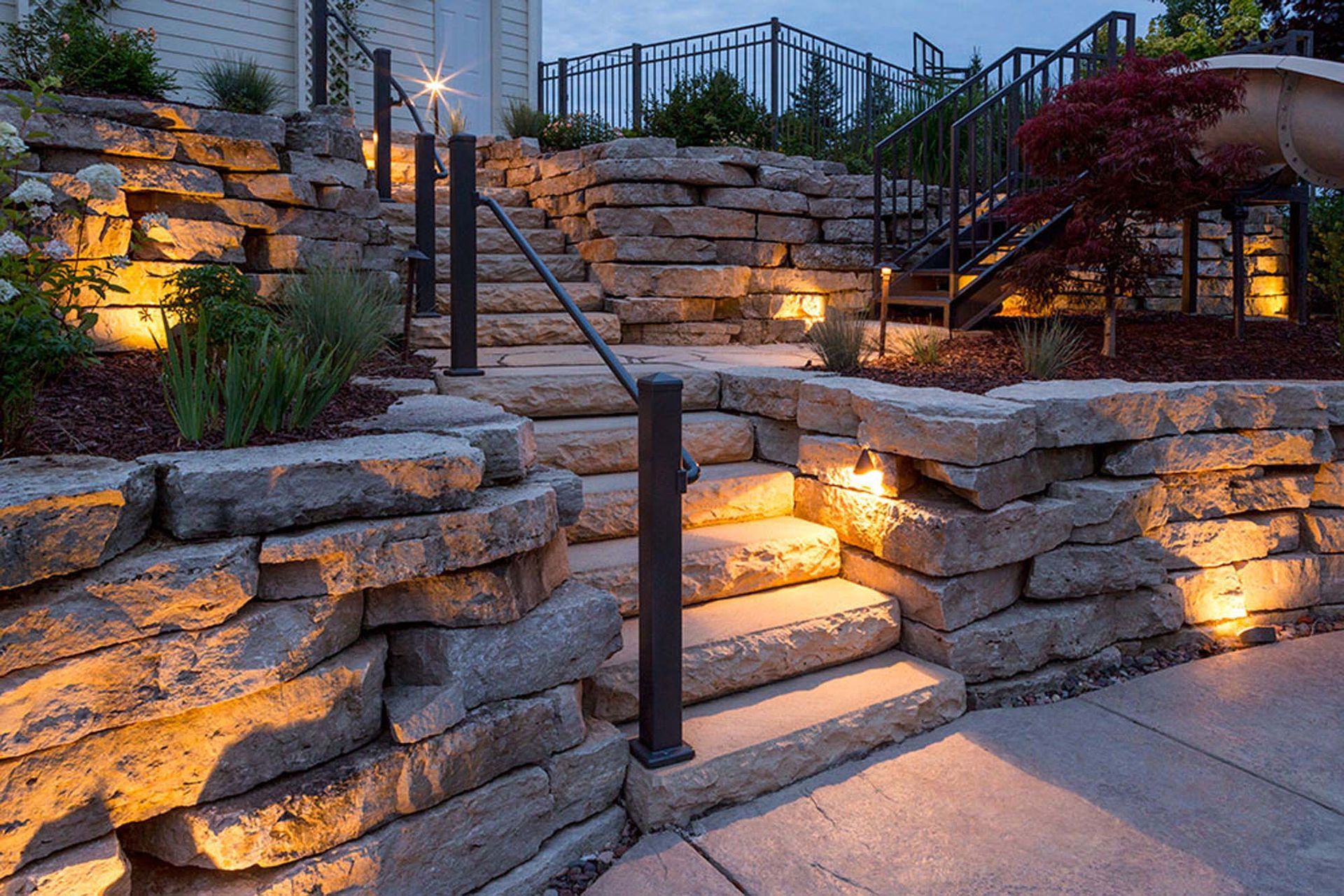 Stone steps with handrails and built-in lighting ascending a terraced landscape at dusk.