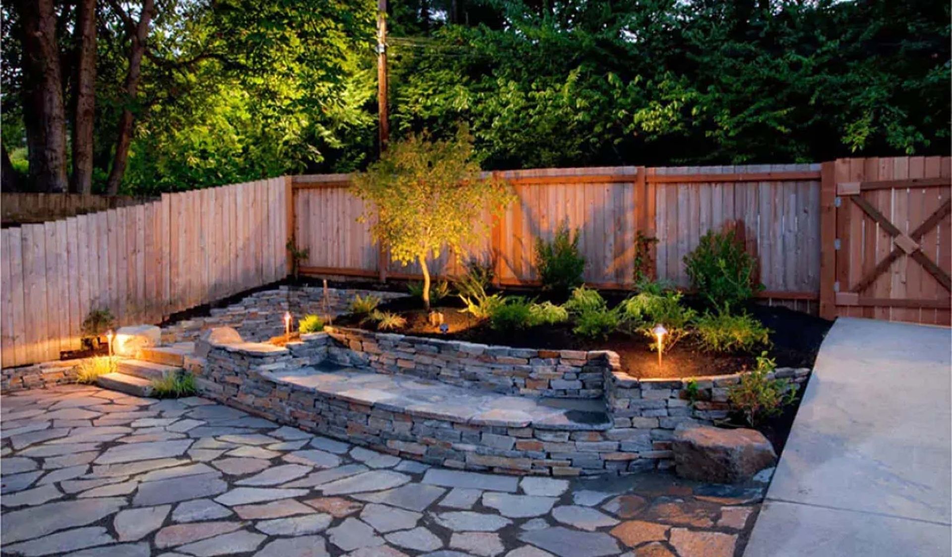 Backyard patio with stone path, low stone wall, lit landscaping, and wooden fence.