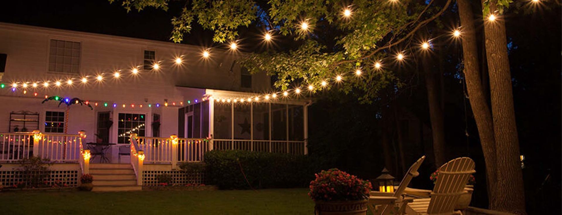 Backyard at night, illuminated by string lights. Deck, house, and trees are visible, with two chairs set up.