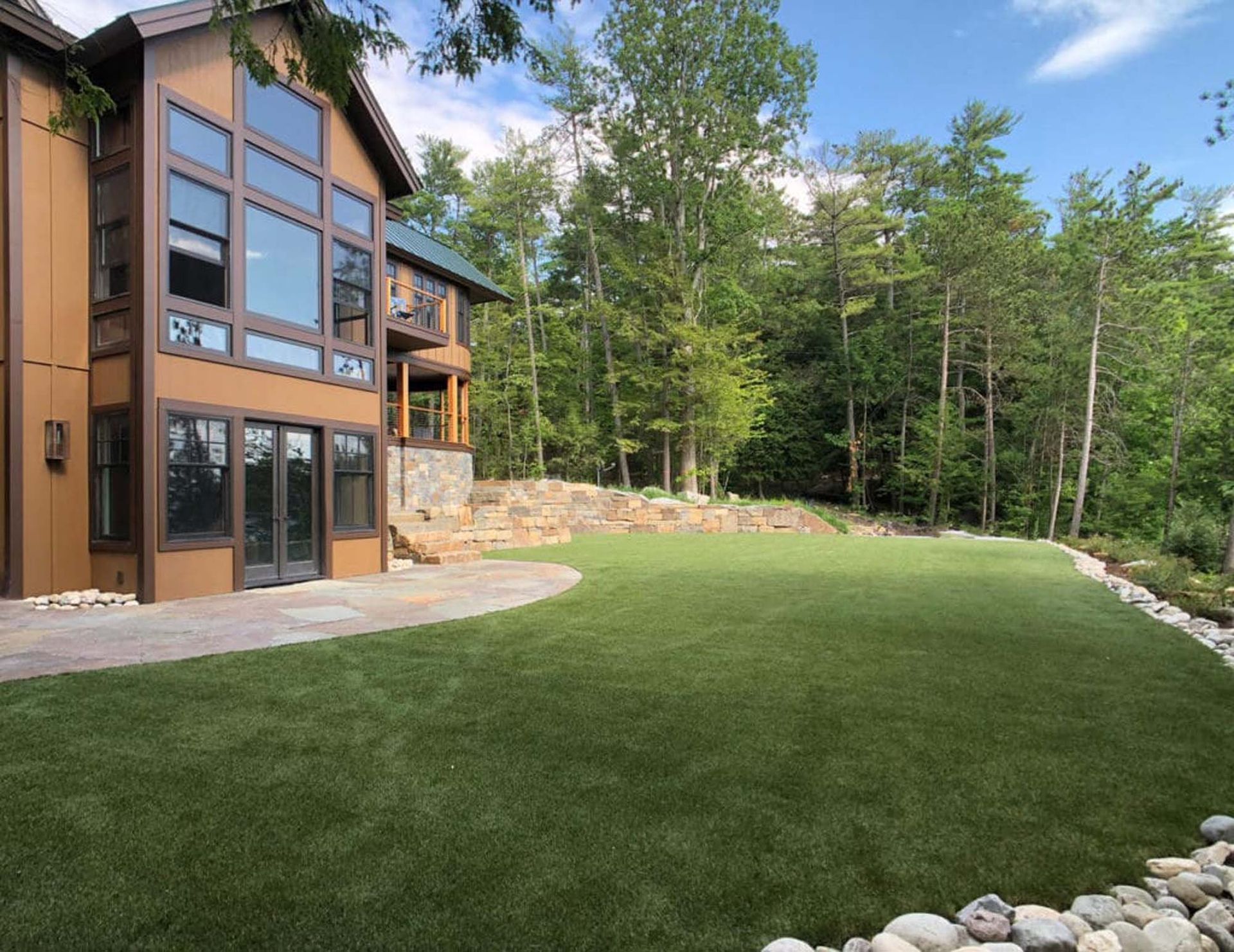 Modern brown house with large windows, grassy lawn, and bordering rocks, set against a forest.