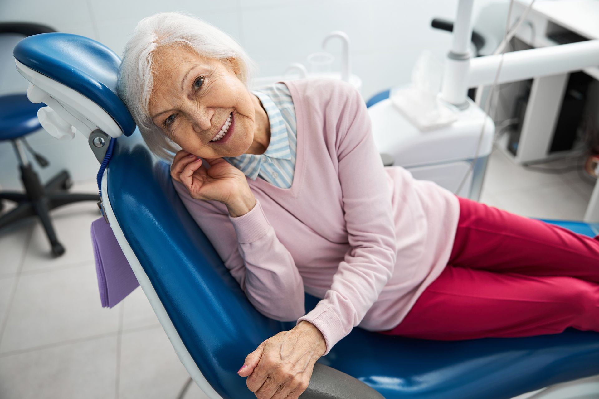 A woman in a dental chair smiles, touching her right cheek after denture repair.