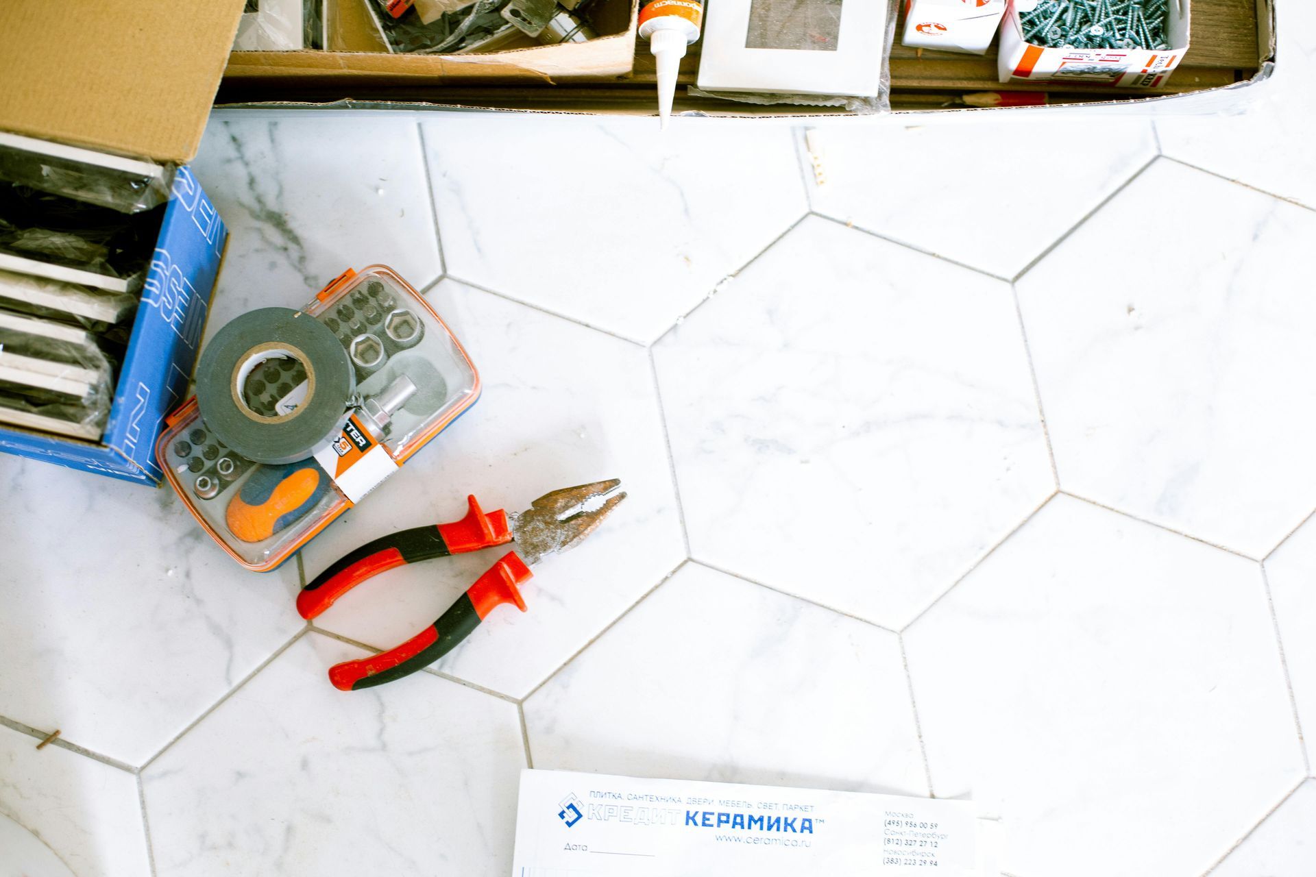 A pair of pliers is sitting on a tiled floor next to a box of tape.