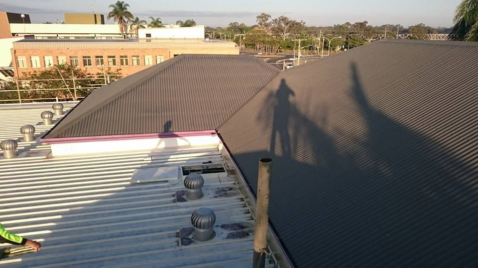 A  Person on A Corrugated Metal Roof, with Buildings — Kaplan Roofing in Walkervale, QLD