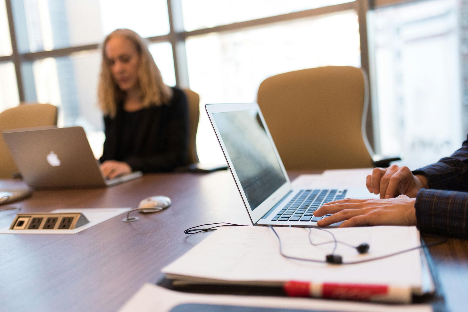 Two people work on laptops at a conference table, with headphones and papers visible in the foreground.