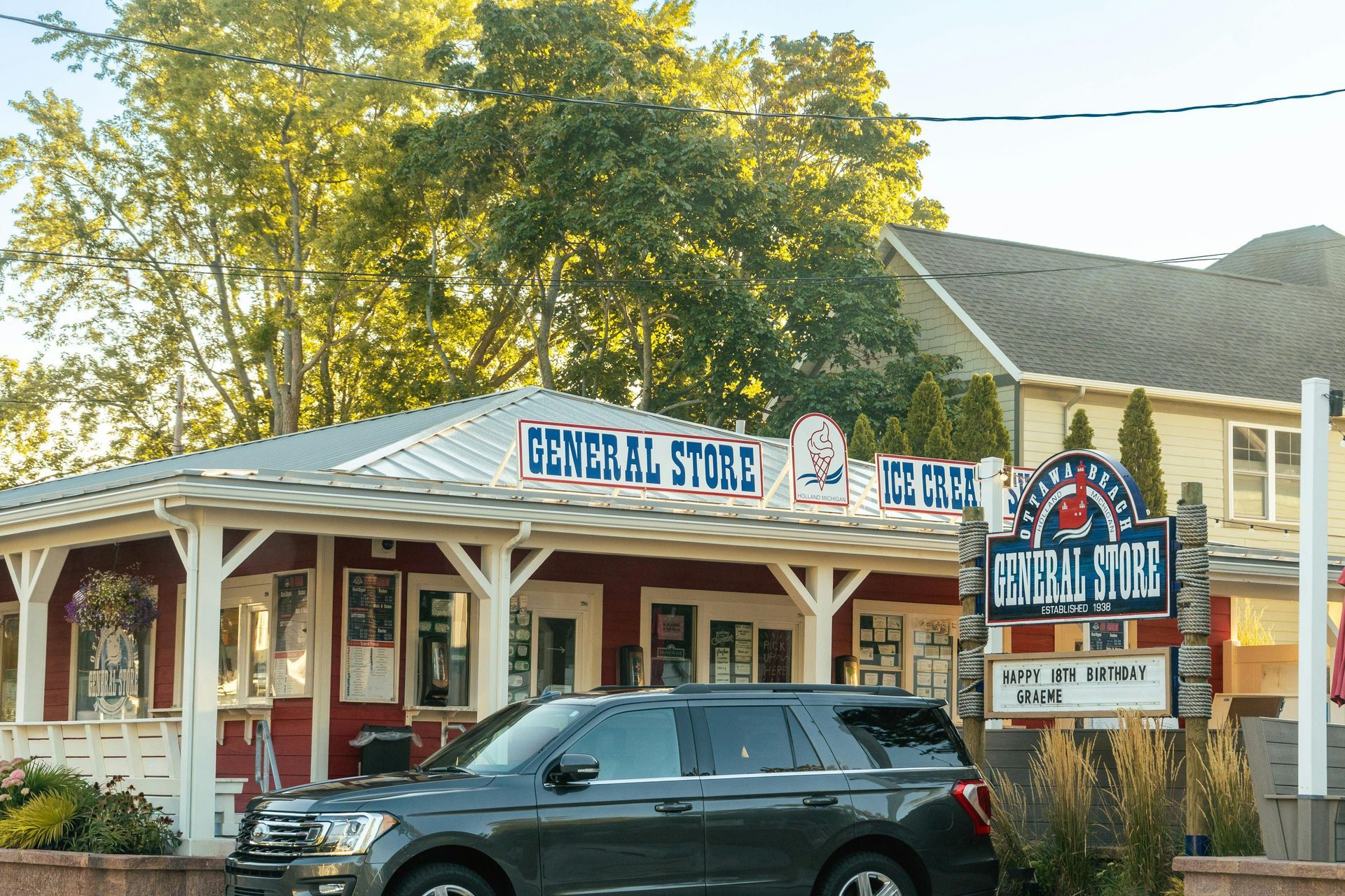 A red general store with white trim and a wrap-around porch, featuring an ice cream sign and a car parked in front.
