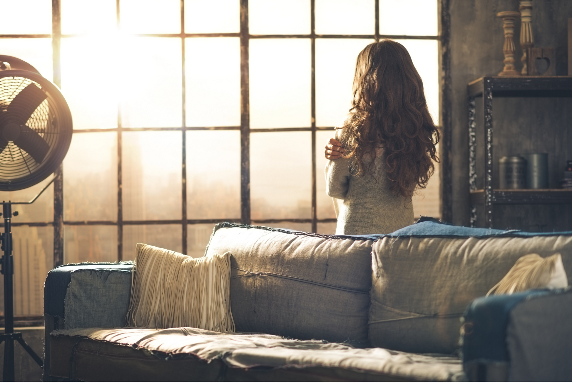 Woman looking out a large window, standing by couch in a room bathed in sunlight.