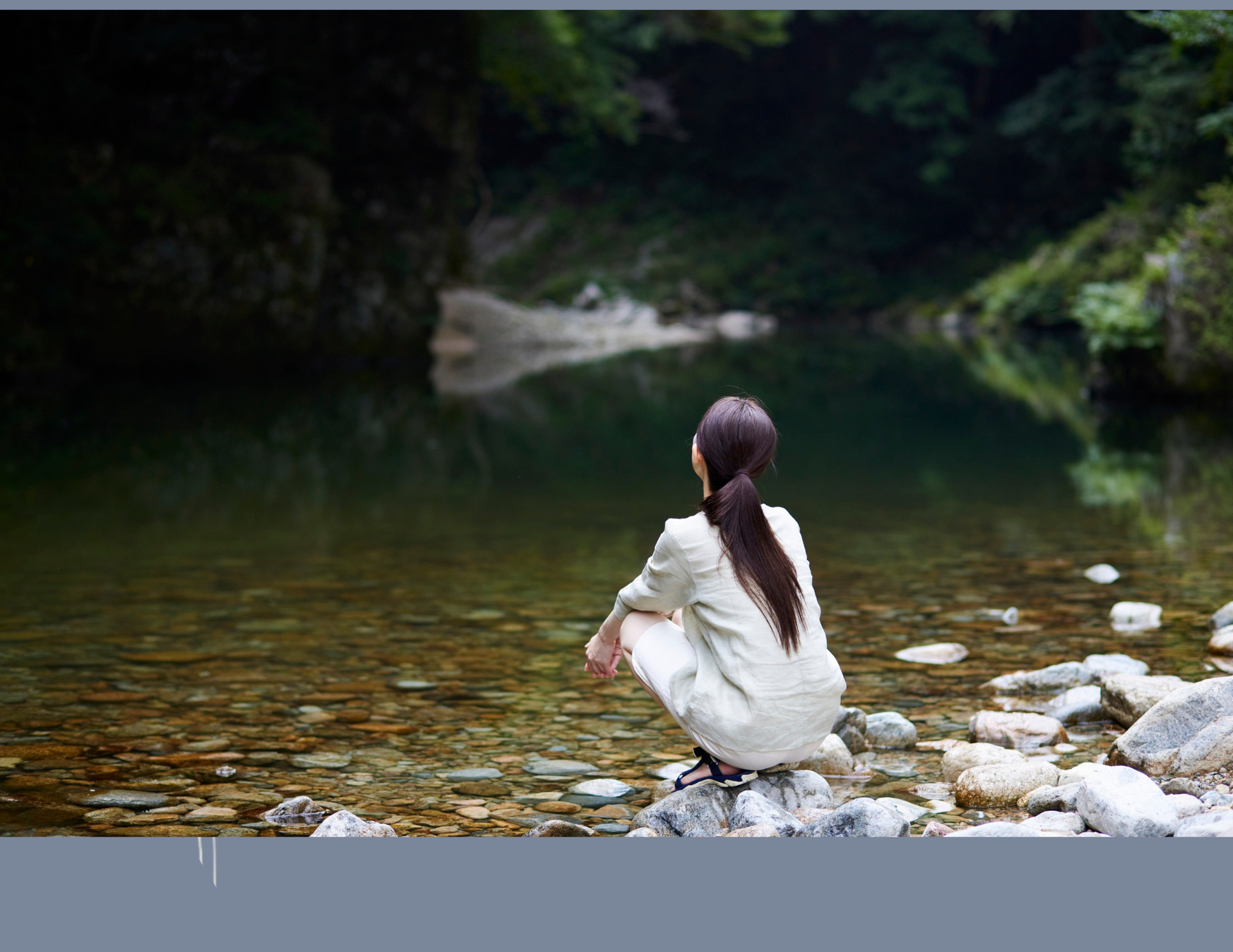 Woman squatting by a calm river, looking at the water. Green trees and rocks surround.