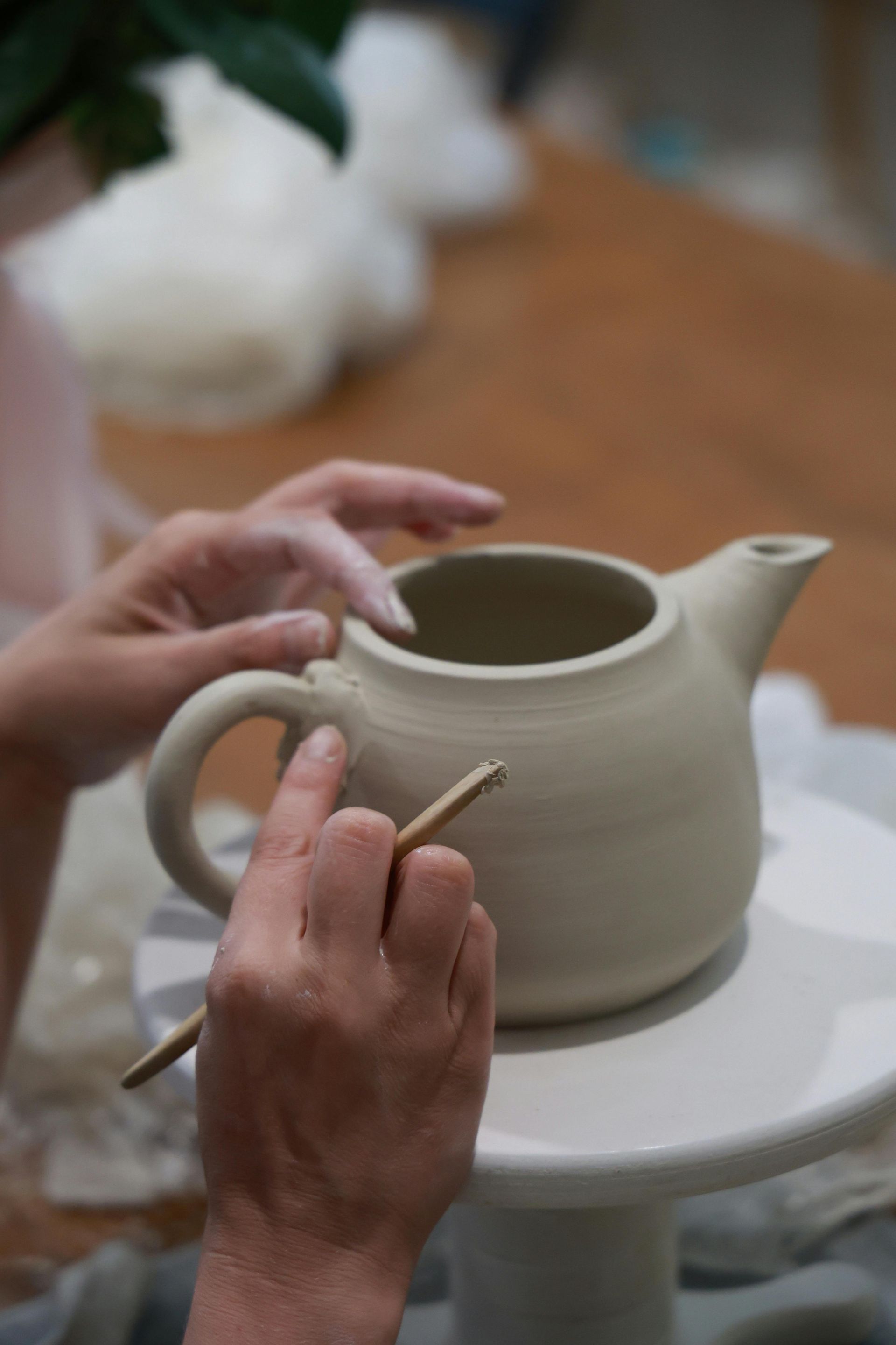 Hands shaping a ceramic teapot with a tool. Light-colored clay, neutral background.