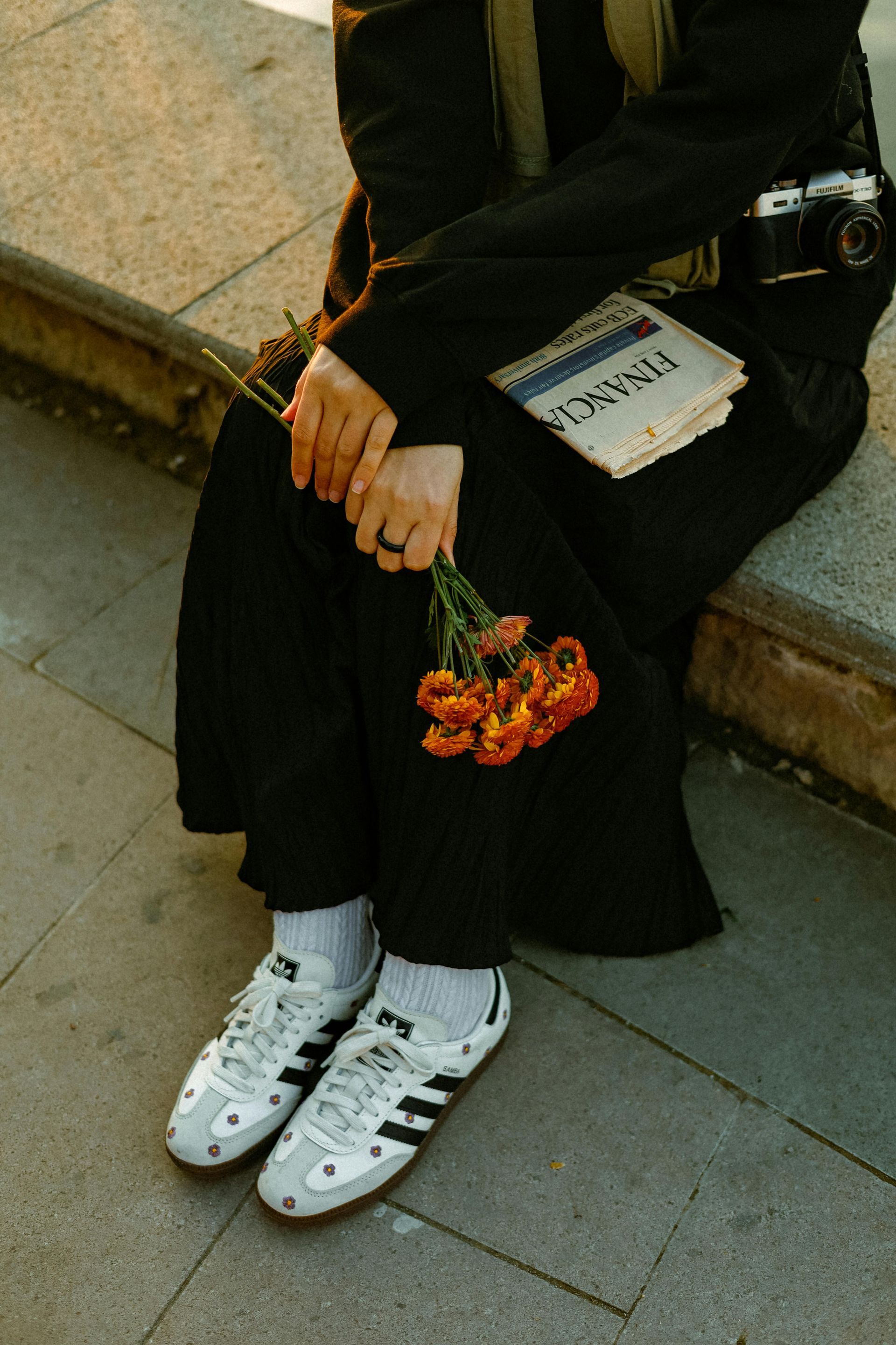 Person in black outfit, holding flowers and newspaper, wearing white sneakers, sitting on a concrete ledge.