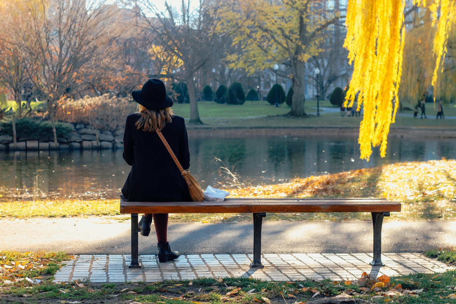 Woman in black coat and hat sits on a bench, facing a lake in autumn.