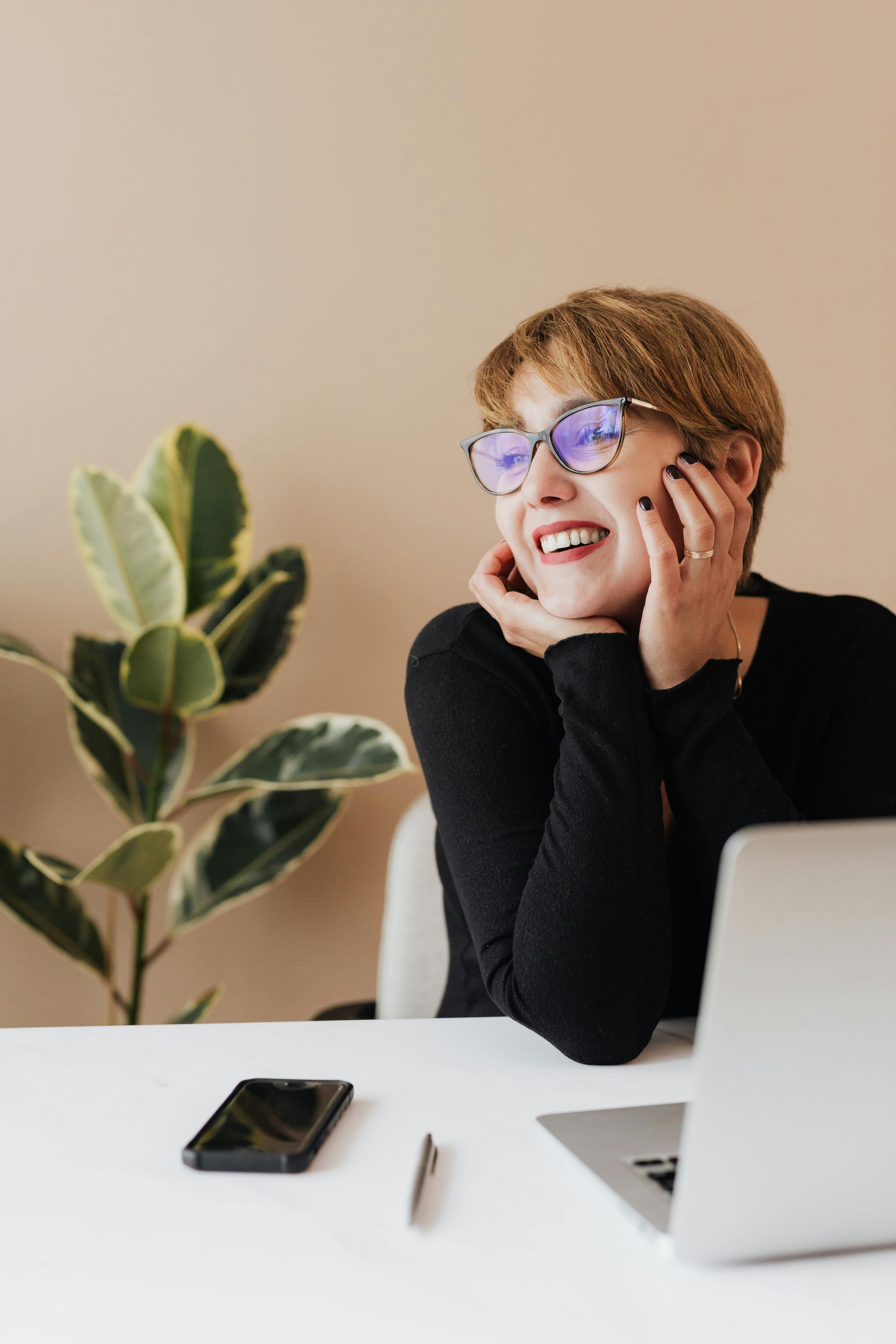 Woman in glasses smiles, resting chin on hands at desk with laptop and phone; plant beside.