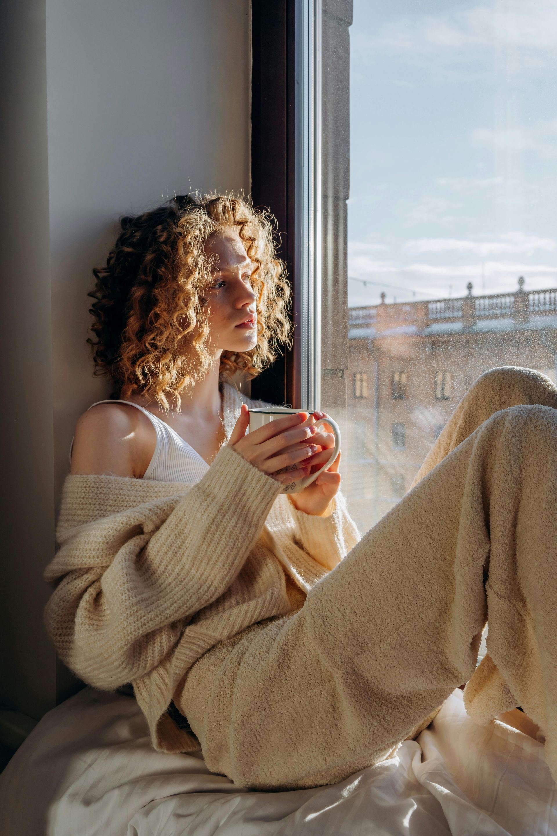 Woman with curly hair sits by window, holding mug, wearing beige sweater.