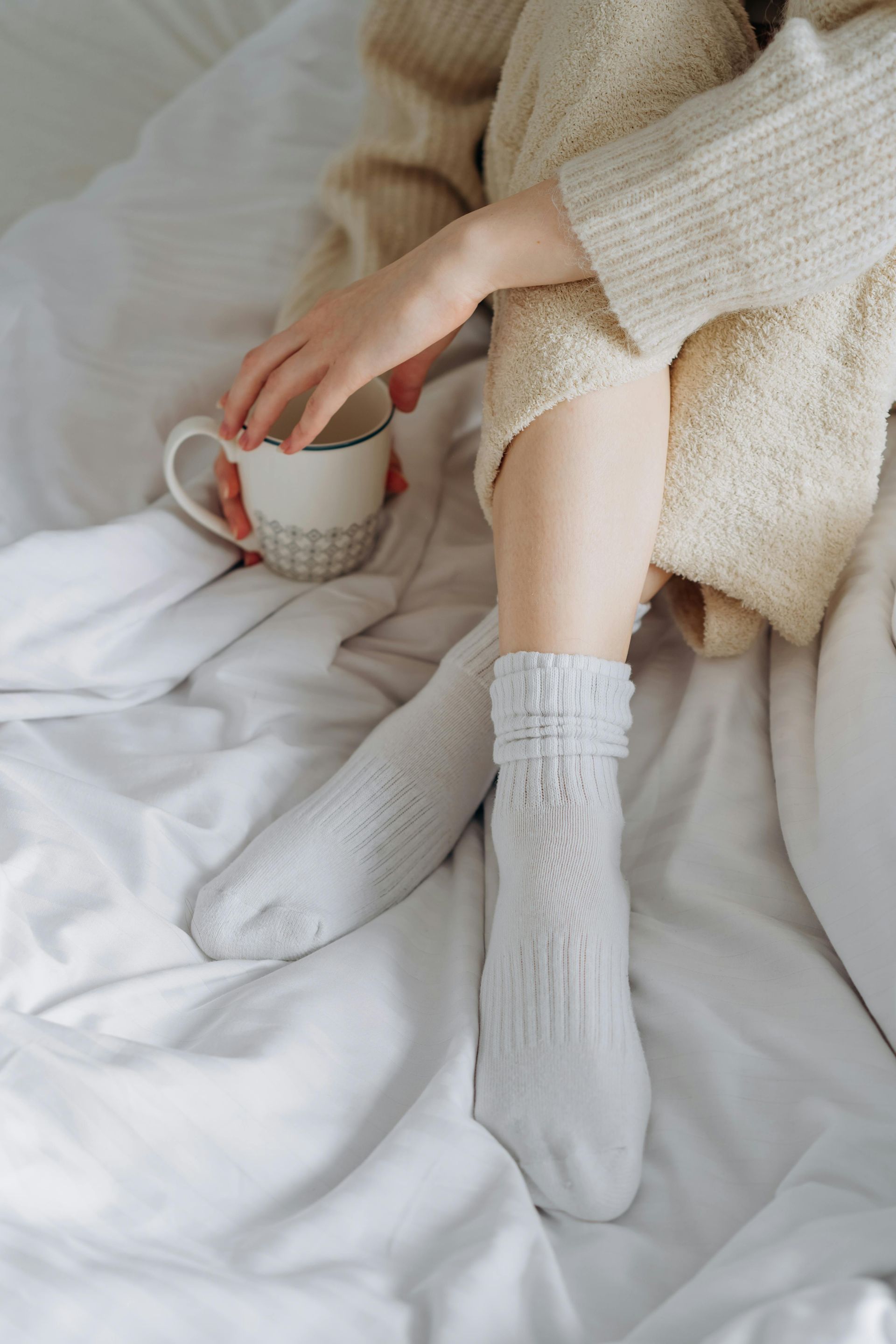 Person in a cream sweater and white socks holding a mug in bed, surrounded by white sheets.