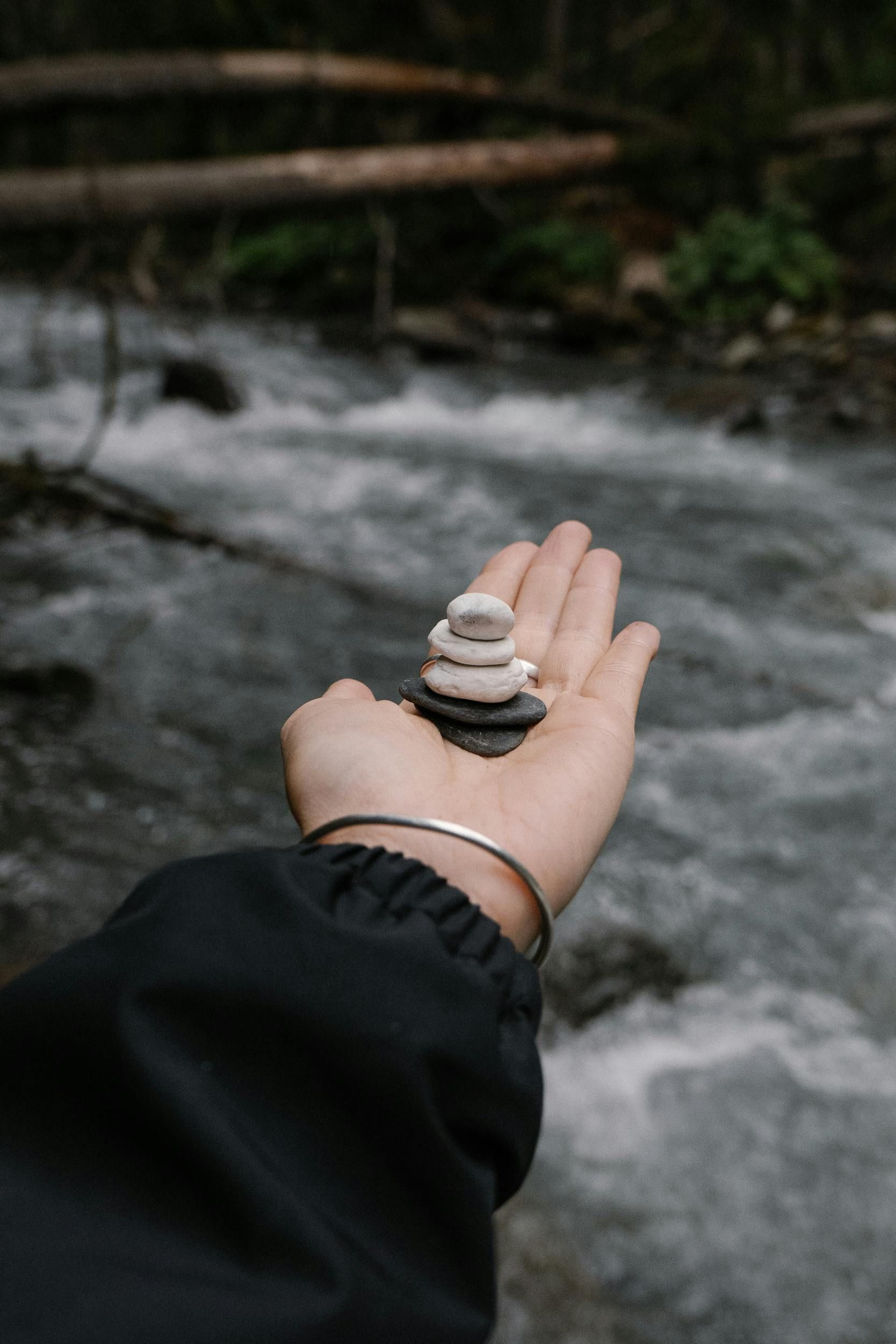 Hand holding a stack of balanced stones, near a flowing stream in a natural setting.