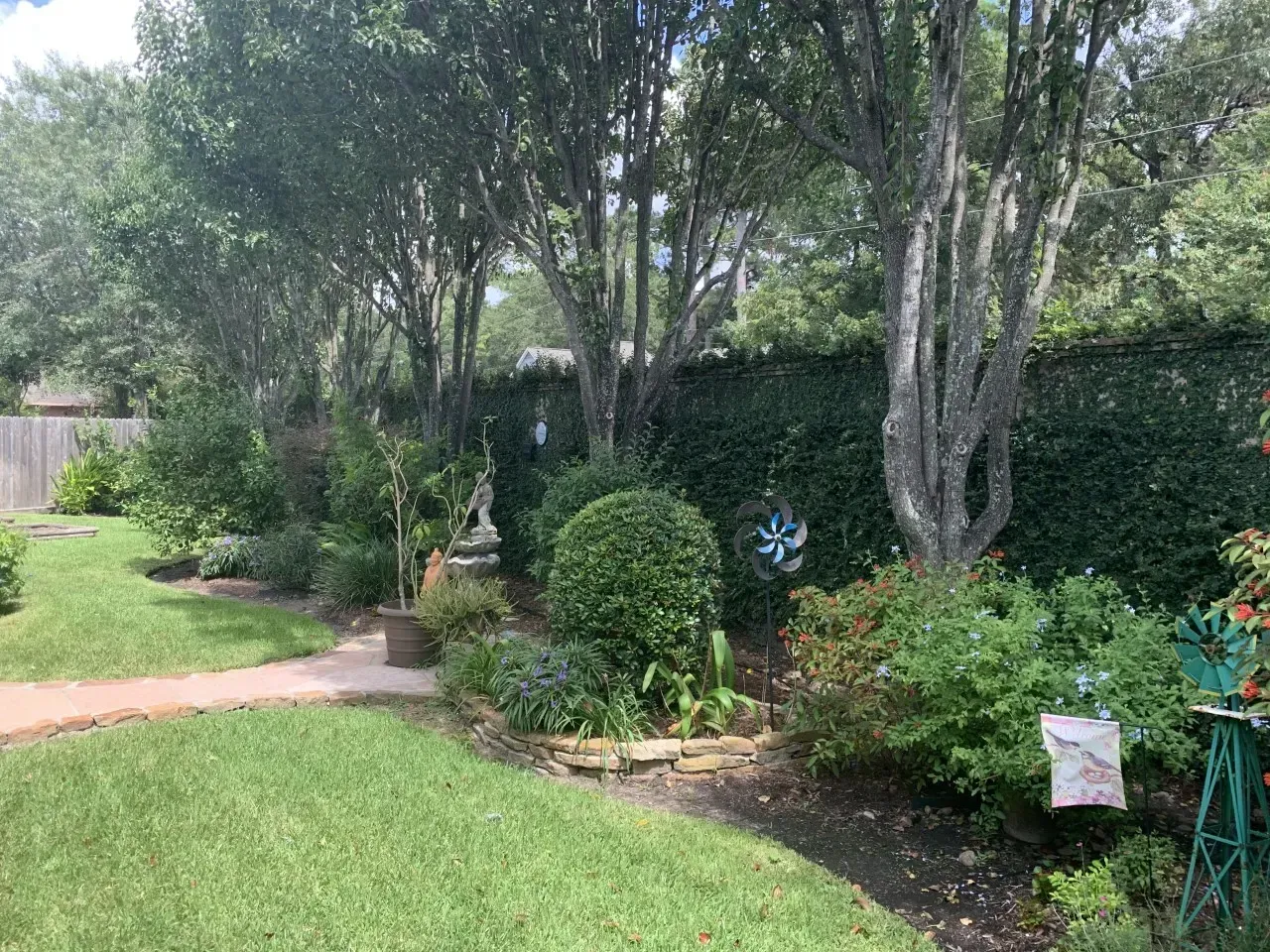 Lush green garden with manicured shrubs, walkway, and tall trees along a fence.