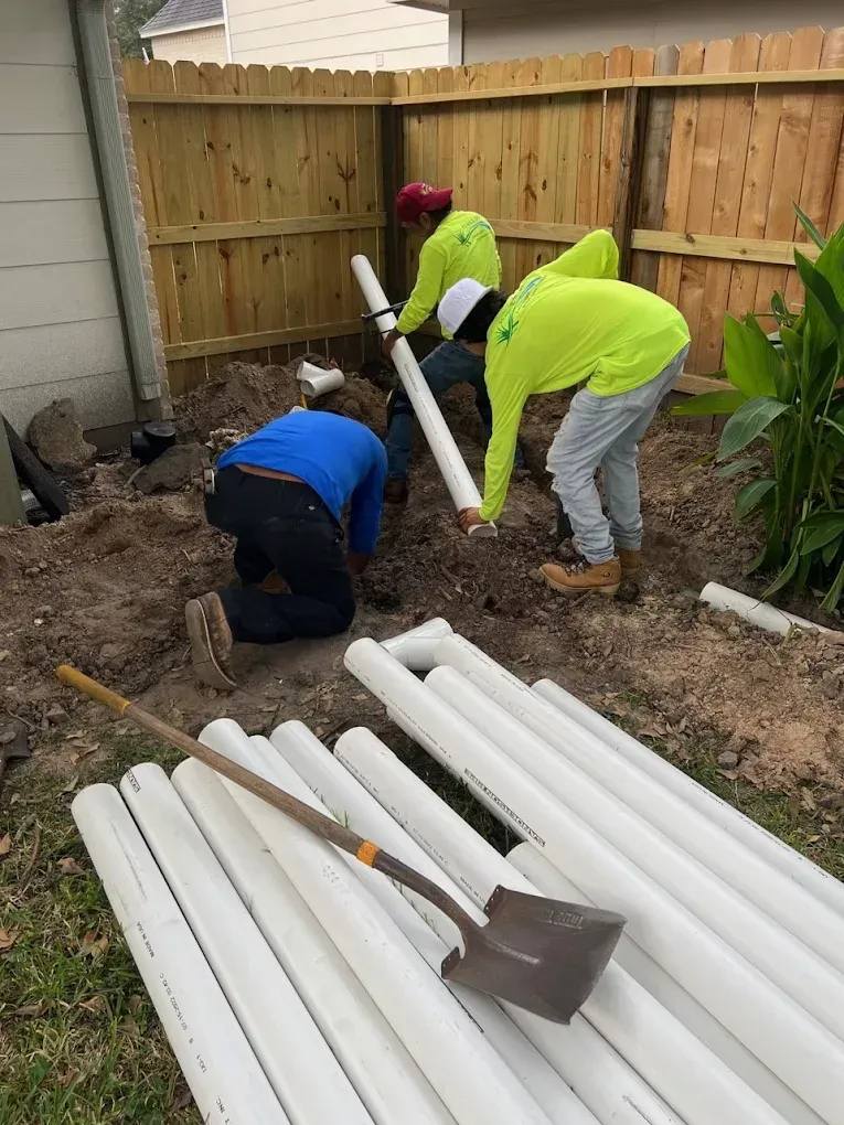 Three workers installing white PVC pipes in a backyard. A shovel and more pipes are in the foreground.