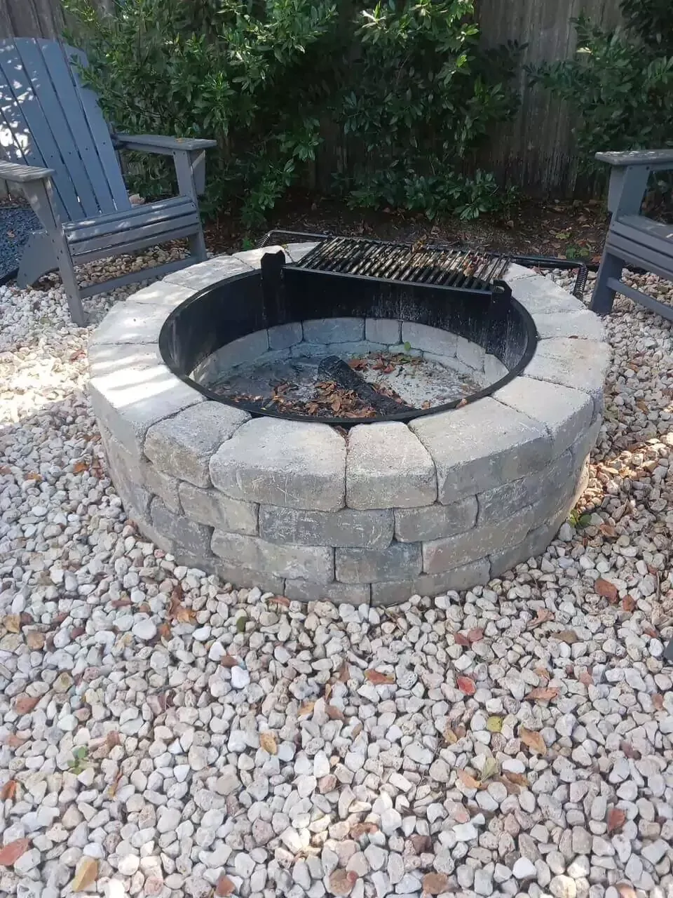 Stone fire pit with a metal grate, surrounded by gravel and two gray chairs.