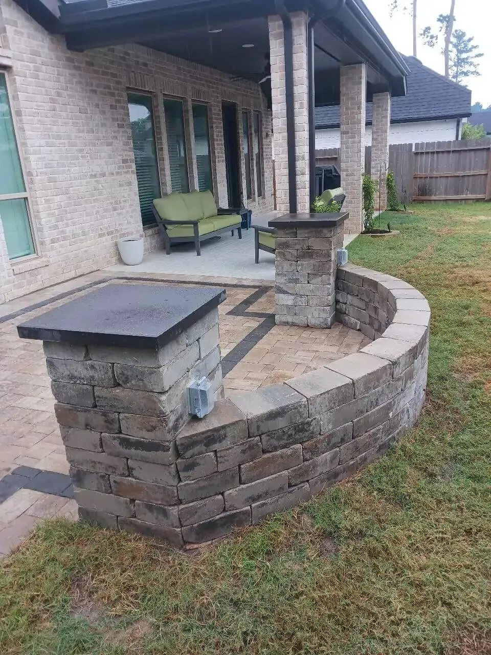 Stone patio with curved retaining wall and brick pillars near a house with green furniture.
