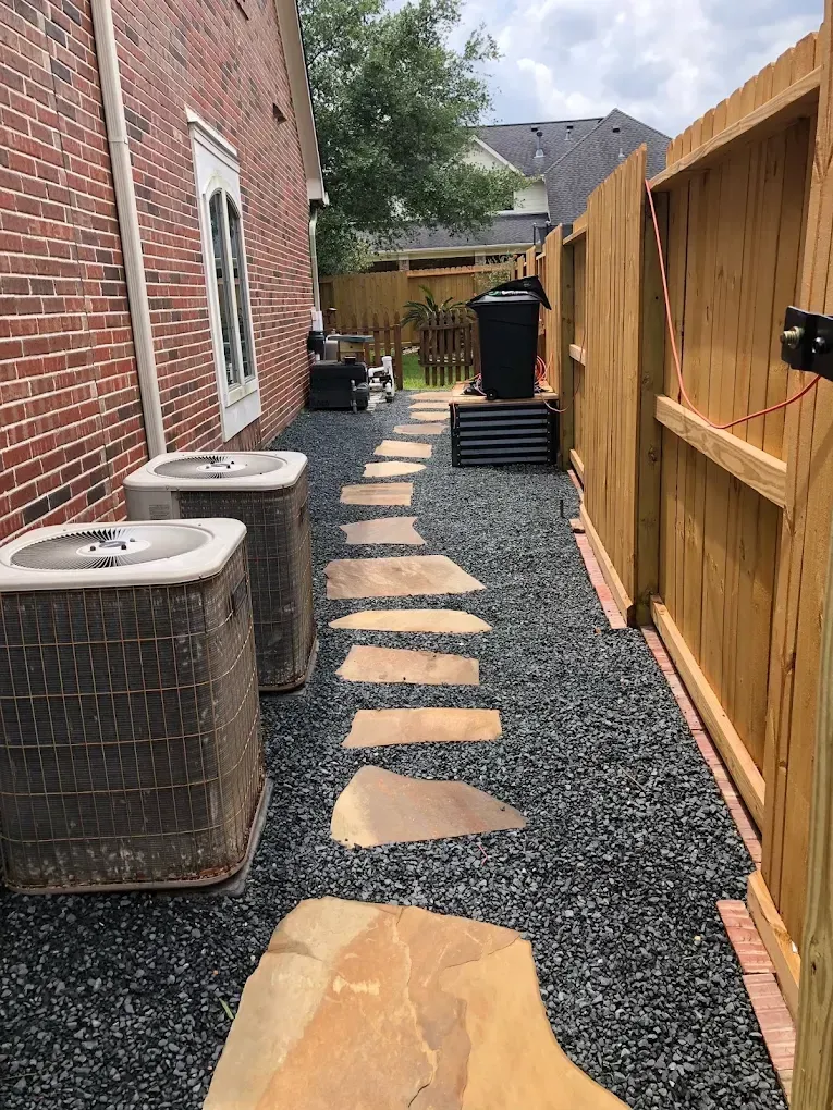 Narrow outdoor path with stepping stones, gravel, air conditioning units, and a wooden fence.