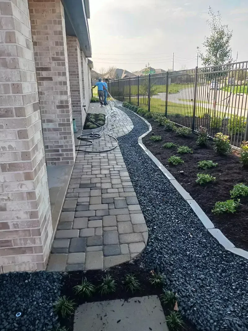 Brick walkway next to gravel path lined with plants, alongside a building and fence; person in distance.