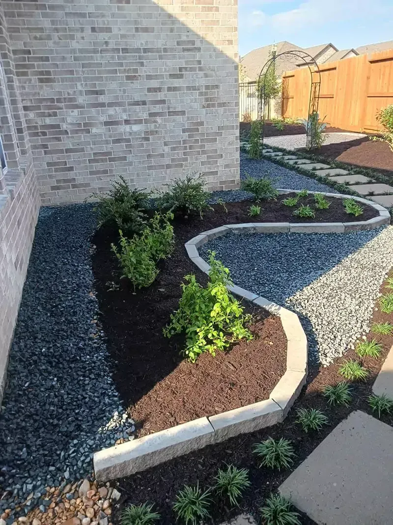 Landscaped yard with winding stone border, plants, mulch, and dark gravel.