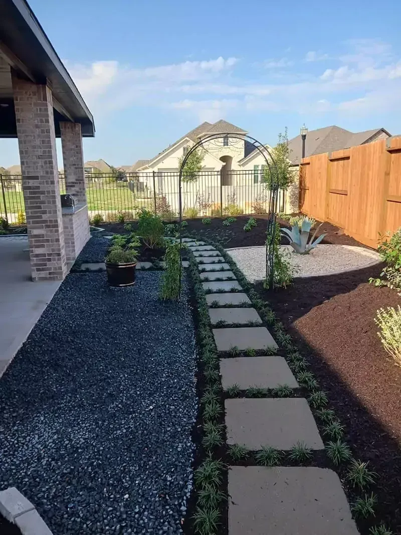 Stone path through a landscaped backyard with dark gravel, dark mulch, and arch.