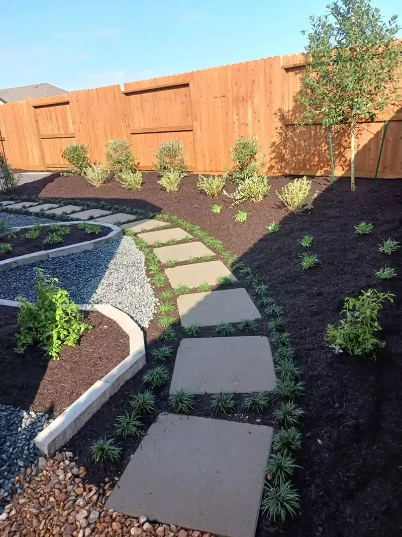 Stone path winds through landscaped backyard with mulch beds, plants, and wooden fence.