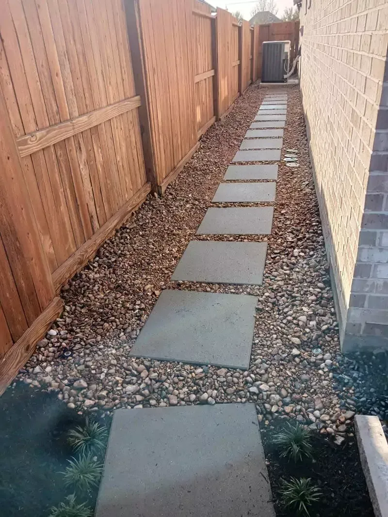 Stone pathway along a wooden fence and brick wall, lined with gravel.