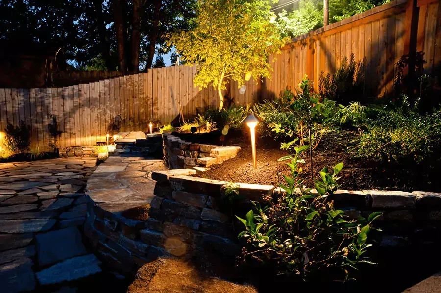 Stone path and tiered garden illuminated by lights at night, next to a wooden fence.
