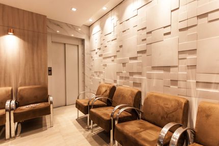 Área de espera moderna com cadeiras marrons dispostas em fila contra uma parede branca texturizada, ao lado de um elevador.