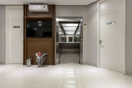 Hall de entrada moderno com uma TV na parede revestida de madeira, um elevador aberto e portas brancas em ambos os lados.