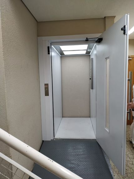 Um elevador aberto branco com luz interna e piso cinza escuro, localizado em um corredor interno de um edifício.
