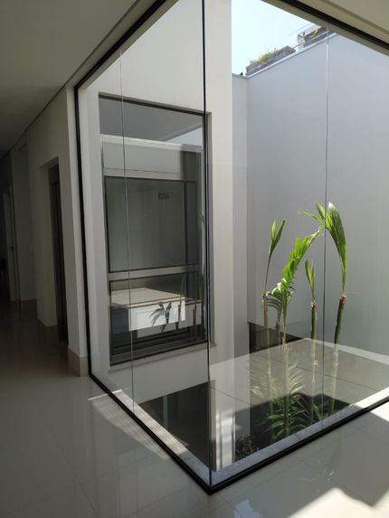 Pátio interno com paredes de vidro, plantas verdes altas e uma parede branca, visível de um corredor com piso de azulejos.