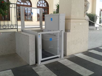 Plataforma elevatória vertical para cadeiras de rodas, de cor cinza claro e guarda-corpos de vidro, na entrada de um prédio.
