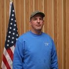 Man in blue sweatshirt and cap standing before a U.S. flag and wood-paneled wall