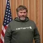 Man standing indoors in front of a U.S. flag, wearing a green Carhartt sweatshirt.