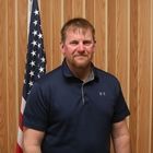 Man standing indoors before wood paneling and a U.S. flag, wearing a navy polo shirt.