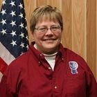 Smiling person in a red shirt standing before an American flag and wood paneling