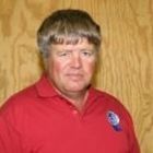 Man in a red polo shirt standing against a wood-paneled wall, wearing a small emblem badge
