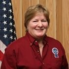 Smiling person in a red shirt standing before a wood-paneled wall and U.S. flag