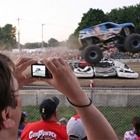 Monster truck jumps over crushed cars at a dirt track while spectators watch and record on phones