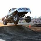 Blue off-road truck airborne over a dirt track jump, with smoke trailing behind.