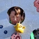 Child peering through a cutout in a painted board, with a small yellow plush toy below.